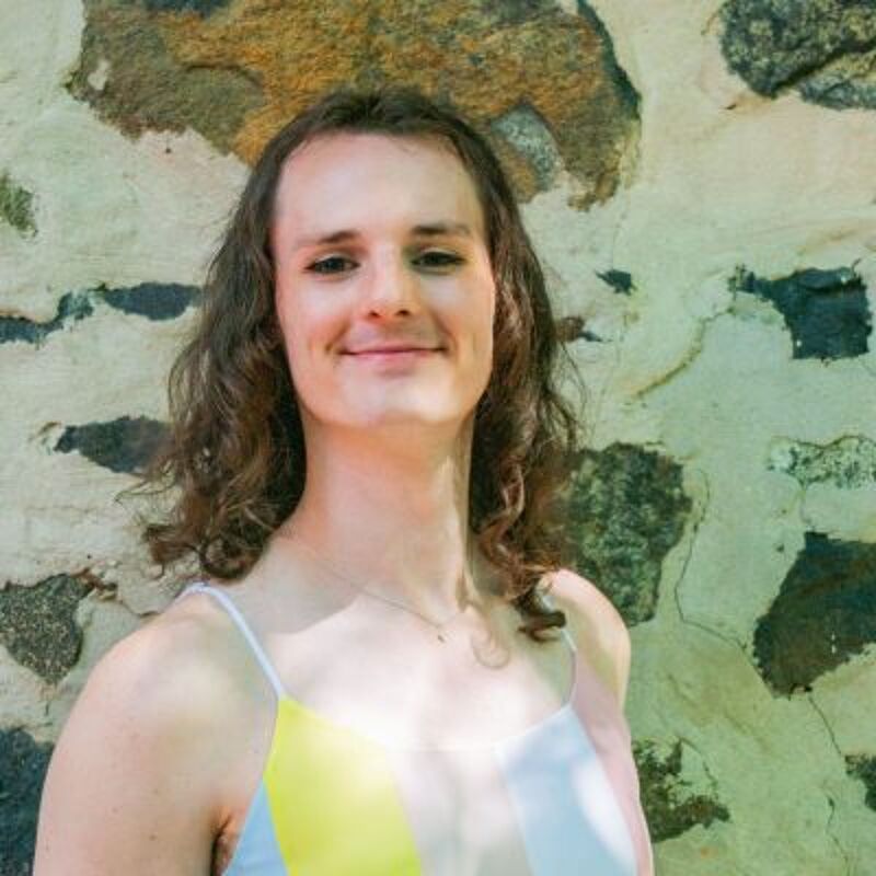 The image shows a person with long, wavy brown hair, fair skin, and a gentle smile. They are wearing a light-colored tank top with yellow and white stripes. The background is a stone wall with various shades of gray and brown. The lighting is soft and natural, highlighting the person's features and creating a warm, inviting atmosphere.